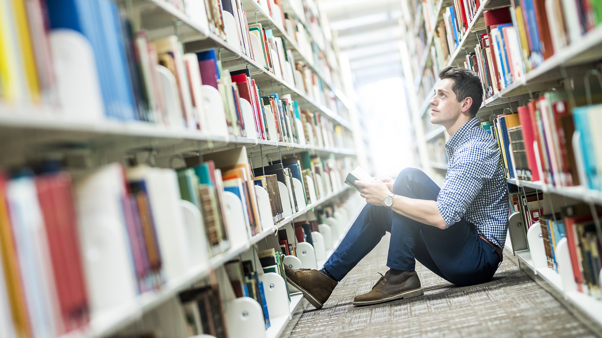 Male student sits on floor between library bookshelves