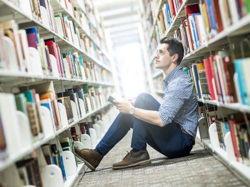 Male student sits on floor between library bookshelves